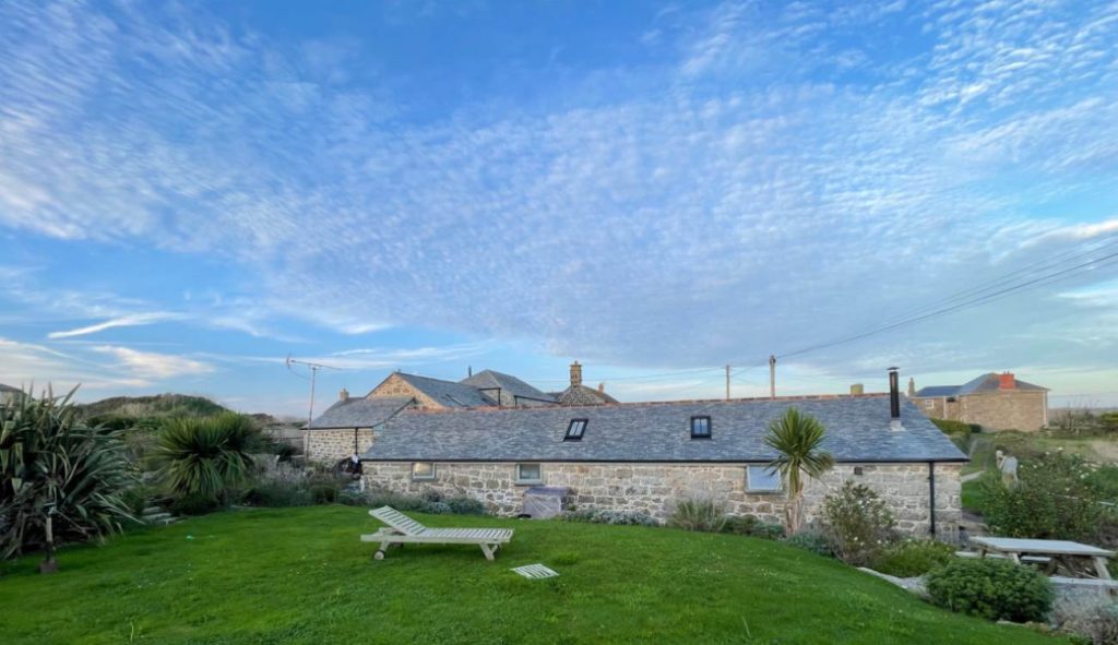 Photo of Trengothal Barn self catering holiday retreat, west of Penzance near Land's End, showing beautiful landscaped garden, side of granite barn and blue sky.